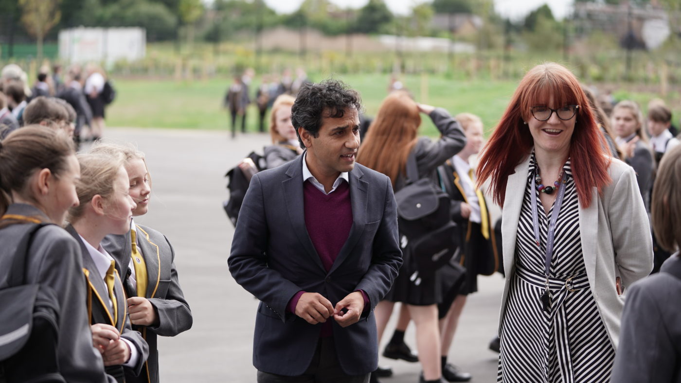 A group of Year 7 students are seen standing on the academy grounds conversing with the Principal and a male member of staff.