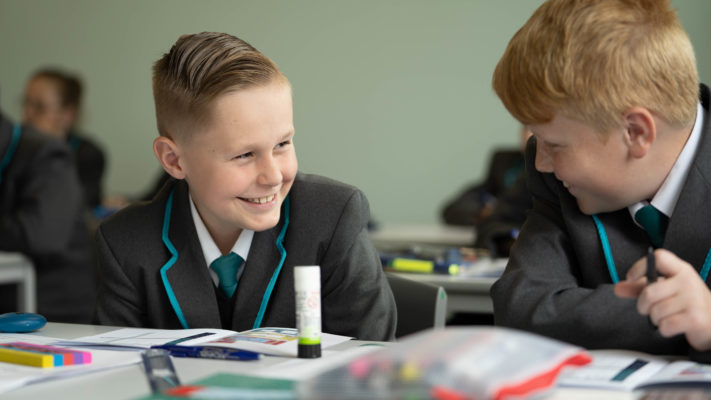 Two male Year 7 students are seen sitting at a desk smiling at each other.