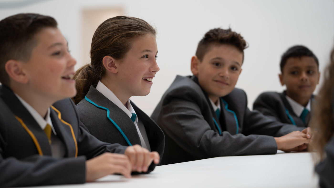 Four students are seen sitting at a desk, smiling for the camera whilst wearing their school uniform.