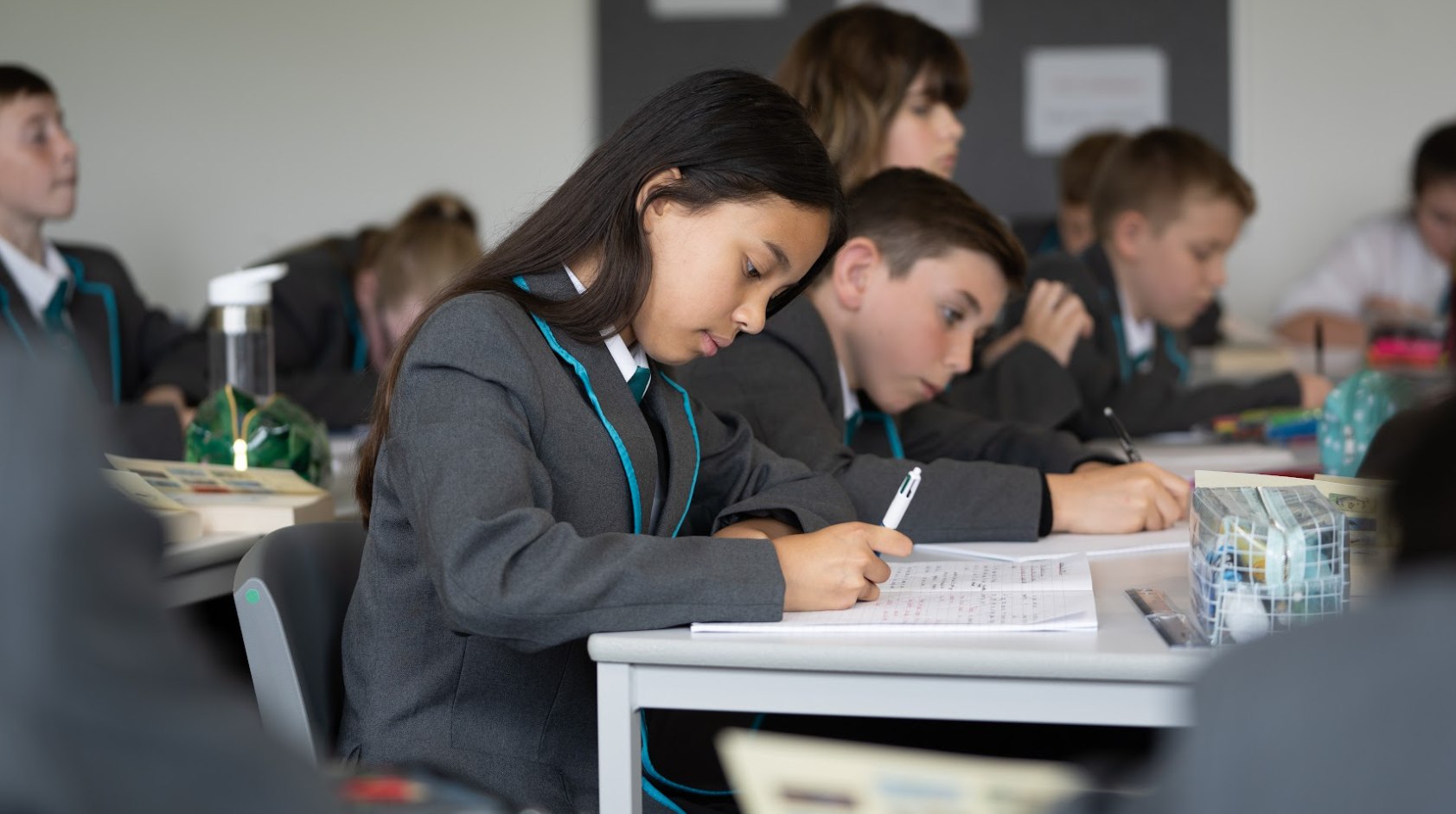 A group of students are seen sitting at their desks in a classroom, writing in their exercise books and wearing their school uniform.