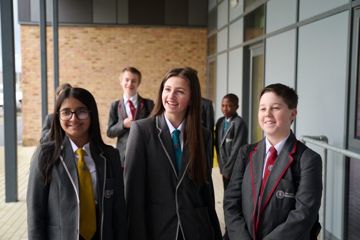 3 students stood outside the school smiling with 4 students in the background also smiling