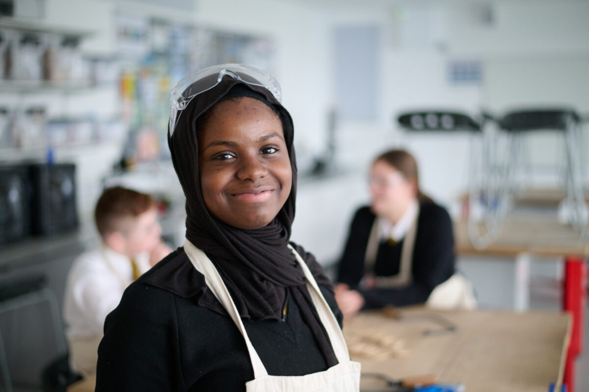 A student in an apron and goggles on her head, smiling for a photo