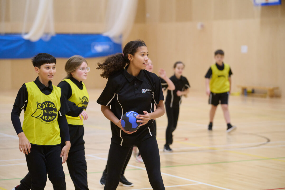 A team of students in a gym in PE kits, with a girl in the foreground holding a ball