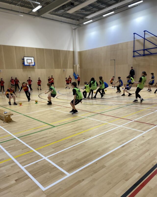 Lots of children in a sports hall playing dodgeball in green or red tabards